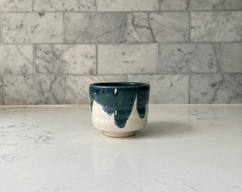 May include: A small, white ceramic teacup with a dark blue glaze that drips down the sides. The cup has a rounded shape and a slightly flared lip.