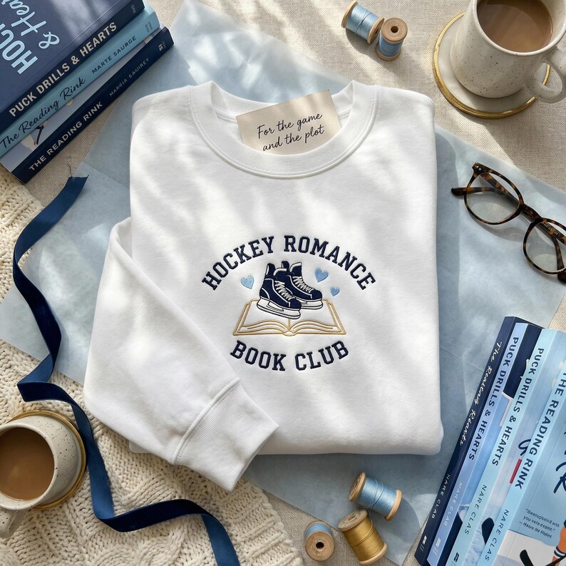 May include: White sweatshirt with the text "HOCKEY ROMANCE BOOK CLUB" in navy blue. The design features hockey skates over an open book, with the phrase "For the game and the plot" on a tag. The sweatshirt is surrounded by books and sewing supplies.
