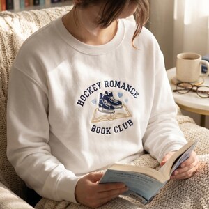May include: White sweatshirt with the text "HOCKEY ROMANCE BOOK CLUB" in navy blue. The design includes hockey skates and hearts. The person is reading a book while sitting in a chair.