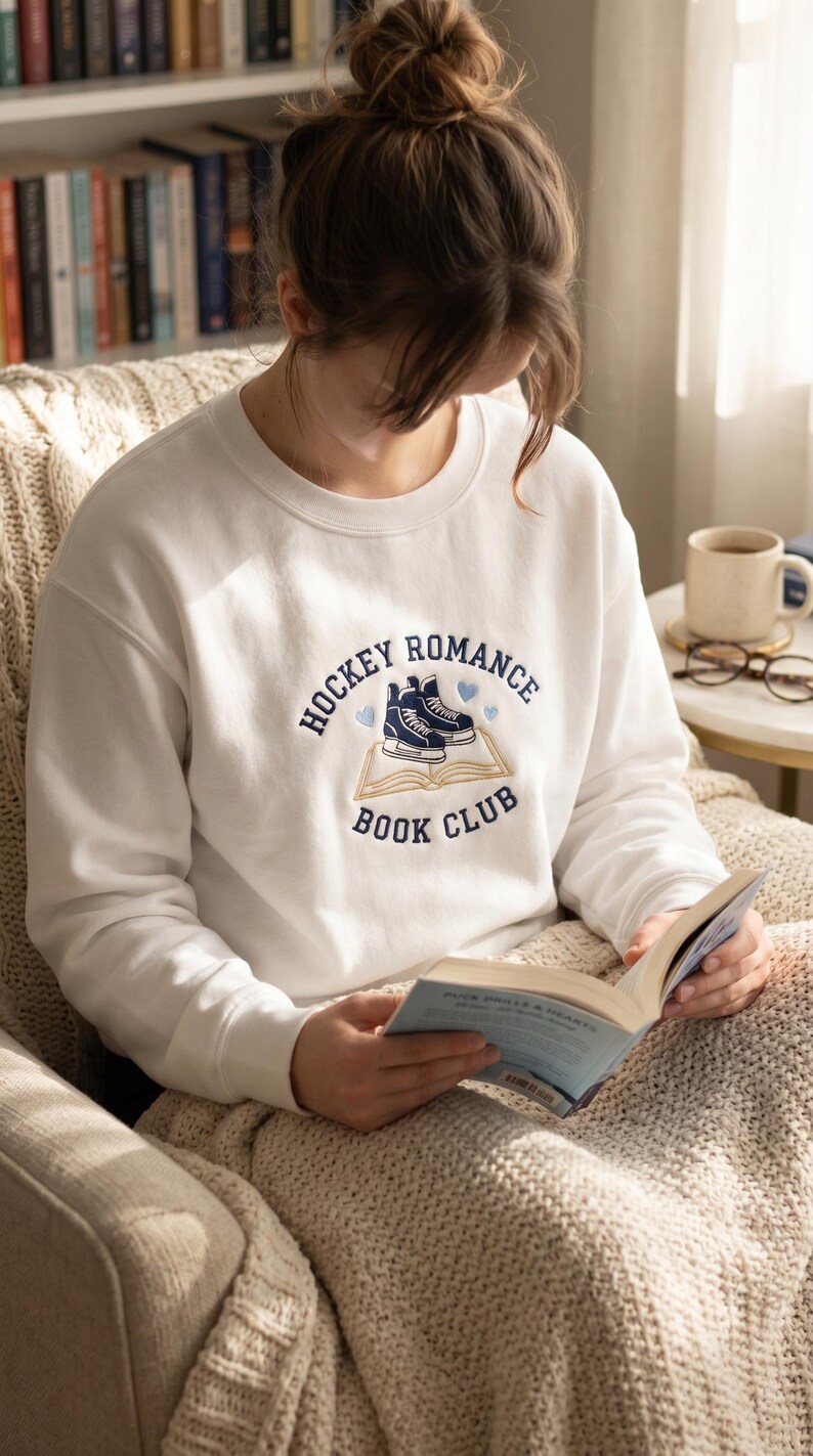 May include: White sweatshirt with the text "HOCKEY ROMANCE BOOK CLUB" in navy blue. The design includes hockey skates and hearts. The person is reading a book while sitting in a chair.