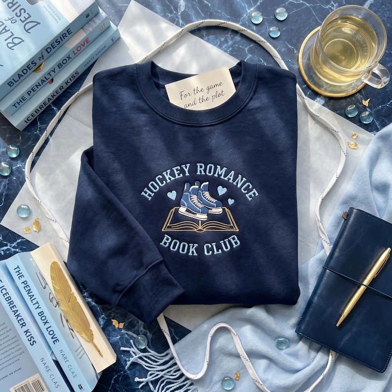 May include: Navy blue sweatshirt with the text "Hockey Romance Book Club" and an illustration of ice skates on an open book. The text "For the game and the plot" is on a small tag. Books and a journal are in the background.