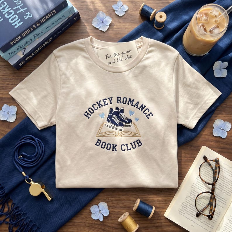 May include: Cream-colored t-shirt with the text "HOCKEY ROMANCE BOOK CLUB" in navy blue. The design features hockey skates on an open book. The shirt also has the text "For the game and the plot".