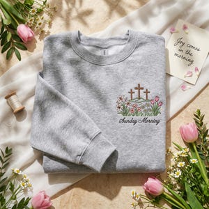 Könnte beinhalten: Ein meliertes graues Sweatshirt mit Rundhalsausschnitt und langen Ärmeln. Auf der Vorderseite befindet sich ein gesticktes Design mit drei Kreuzen auf einem Hügel, umgeben von Blumen, mit dem Text "Sunday Morning" darunter. Eine Karte mit dem Text "Joy comes in the morning" ist ebenfalls sichtbar.