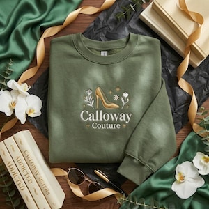 May include: An olive green sweatshirt with an embroidered gold high heel design and the text "Calloway Couture". The sweatshirt is surrounded by books, sunglasses, and flowers, suggesting a fashion or lifestyle theme.