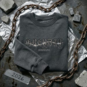 May include: A dark gray sweatshirt with the word "GREYDAY" embroidered in a gothic font, with barbed wire detail. The year "MMXXVI" is below the word. The sweatshirt is on a metallic surface with a rusty chain and concrete pieces.