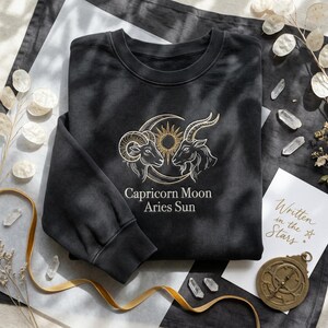 May include: A black sweatshirt featuring an embroidered design of two zodiac signs, a ram and a goat, with a sun and moon. The text "Capricorn Moon Aries Sun" is below the design. A card with the text "Written in the Stars" is also visible.