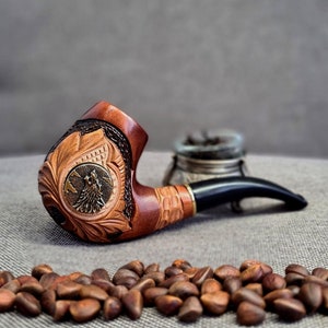 May include: A wooden pipe with a carved design of a wolf's head. The pipe has a black stem and is resting on a surface with pine nuts.