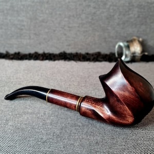 Könnte beinhalten: Eine dunkelbraune Holzpfeife mit einem schwarzen Mundstück und goldenen Akzenten. Die Pfeife hat einen gebogenen Kopf und einen langen Schaft.