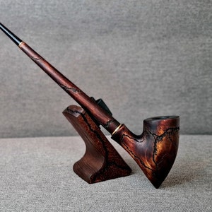 Könnte beinhalten: Eine dunkelbraune Holzpfeife mit einem langen, gebogenen Schaft und einem Kopf mit einem einzigartigen, strukturierten Design. Die Pfeife liegt auf einem Holzständer.