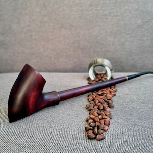 Könnte beinhalten: Eine weinrote Holzpfeife mit schwarzem Mundstück, die auf einer grauen Oberfläche liegt. Ein kleiner silberner Behälter ist umgestürzt und verstreut eine Spur brauner Pinienkerne. Die Pfeife ist ein klassisches Raucherzubehör.