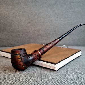 Op de afbeelding: Een bruine houten pijp met een zwarte steel die op een bruin notitieboekje ligt. De pijp heeft een marmerpatroon en een gouden band rond de kom.