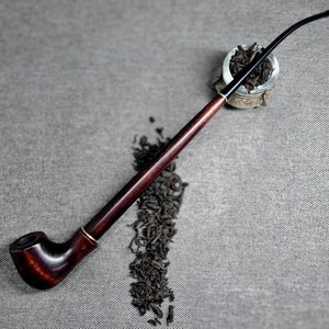 Puede incluir: Una pipa de madera marrón oscuro con una boquilla negra y una cazoleta llena de hojas de té sueltas. La pipa está sobre una superficie gris texturizada.