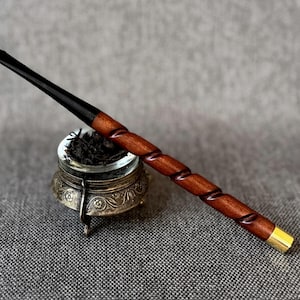 May include: A wooden smoking pipe with a spiral design and a black mouthpiece rests near a small, ornate metal container. The pipe has a dark brown finish with a gold-colored metal band at the end.