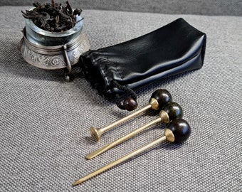 Könnte beinhalten: Drei goldene Teewerkzeuge aus Metall mit schwarzen und grünen Holzgriffen. Die Werkzeuge befinden sich in einer schwarzen Ledertasche. Ein silberner Teekasten mit losen Teeblättern befindet sich im Hintergrund.