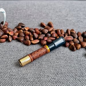 May include: A silver and glass container with a decorative design sits on a gray surface, spilling out brown pine nuts. A wooden and gold cigarette holder with a carved design lies next to the container.