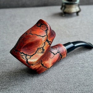 Puede incluir: Una pipa de madera roja y marrón con una boquilla negra. La pipa tiene un patrón único de veta de madera natural.