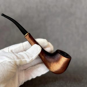 Könnte beinhalten: Eine Tabakpfeife aus Holz mit einem gebogenen schwarzen Mundstück und einem goldfarbenen Band. Der Kopf ist dunkelbraun und der Stiel wird in einer behandschuhten Hand gehalten. Die Pfeife hat ein klassisches, traditionelles Design.