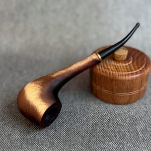 May include: A wooden smoking pipe with a polished brown bowl and a long, slender black stem. The pipe features a gold-coloured metal ring at the junction of the bowl and stem. Beside the pipe sits a small, round, light brown wooden container.