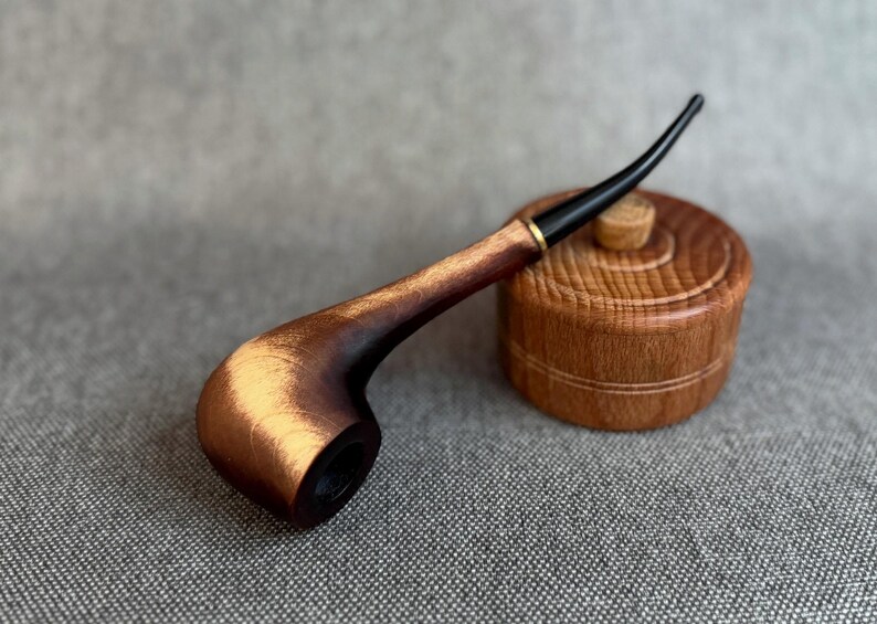 May include: A wooden smoking pipe with a polished brown bowl and a long, slender black stem. The pipe features a gold-coloured metal ring at the junction of the bowl and stem. Beside the pipe sits a small, round, light brown wooden container.