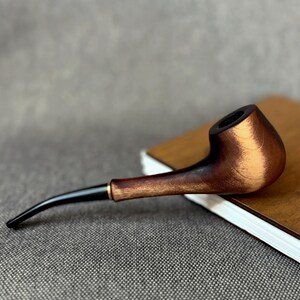 May include: A tobacco pipe featuring a polished wooden bowl with a dark brown and light brown wood grain finish, connected to a slender, curved black stem with a small gold-coloured metal ring at the junction of the stem and the bowl.