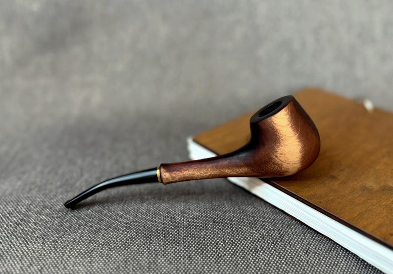 May include: A tobacco pipe featuring a polished wooden bowl with a dark brown and light brown wood grain finish, connected to a slender, curved black stem with a small gold-coloured metal ring at the junction of the stem and the bowl.