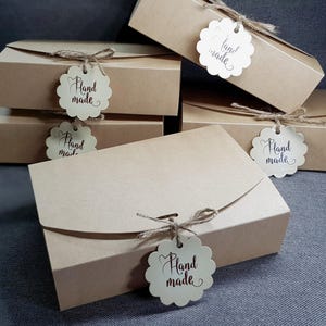 May include: A collection of rectangular light brown cardboard boxes, each featuring a curved flap closure secured with natural jute twine. A scalloped white paper tag with the text Hand made in dark script is tied to each box.