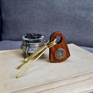 May include: A wooden hair accessory with a circular cutout and a round metal tree emblem, paired with two thin brass hair sticks. Next to it sits a small, ornate silver-toned metal bowl filled with dark, loose-leaf tea or herbs.