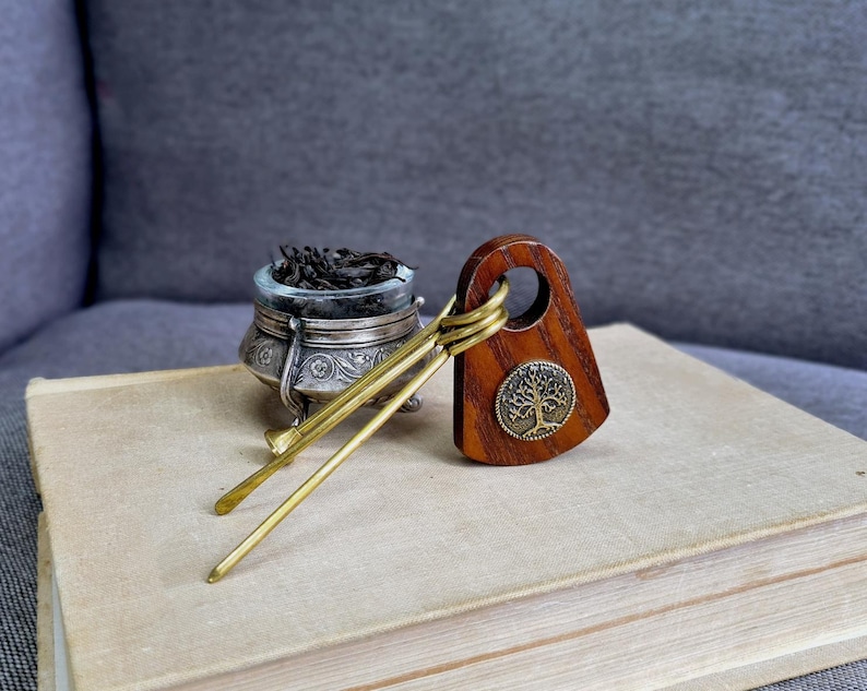 May include: A wooden hair accessory with a circular cutout and a round metal tree emblem, paired with two thin brass hair sticks. Next to it sits a small, ornate silver-toned metal bowl filled with dark, loose-leaf tea or herbs.