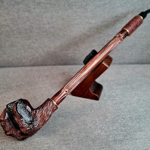 May include: A wooden smoking pipe with a carved bear paw bowl. The pipe is brown and has a natural wood finish. It is displayed on a wooden stand.