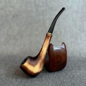 May include: A tobacco pipe crafted from polished brown wood with a prominent grain pattern, featuring a long, curved black stem and a small gold-coloured ring at the junction, resting against a matching curved wooden stand.