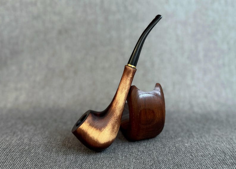 May include: A tobacco pipe crafted from polished brown wood with a prominent grain pattern, featuring a long, curved black stem and a small gold-coloured ring at the junction, resting against a matching curved wooden stand.