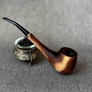 May include: A tobacco pipe with a polished brown wooden bowl and a slender black stem featuring a gold-coloured band. The pipe rests against a small, ornate silver-toned metal container with intricate floral engravings filled with dark tobacco.