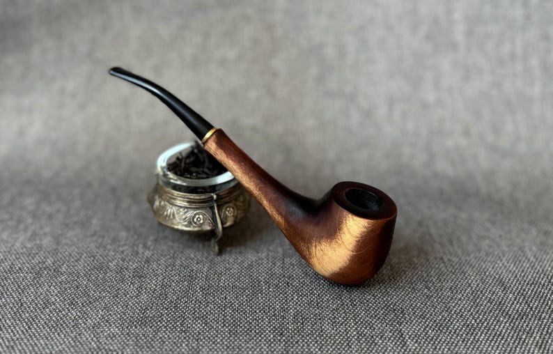 May include: A tobacco pipe with a polished brown wooden bowl and a slender black stem featuring a gold-coloured band. The pipe rests against a small, ornate silver-toned metal container with intricate floral engravings filled with dark tobacco.