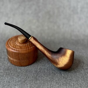 May include: A polished wooden tobacco pipe with a curved black stem and a gold-coloured metal band, resting against a small, round, lidded wooden container featuring a textured, grooved surface and a central knob handle.