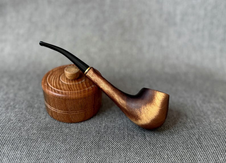 May include: A polished wooden tobacco pipe with a curved black stem and a gold-coloured metal band, resting against a small, round, lidded wooden container featuring a textured, grooved surface and a central knob handle.