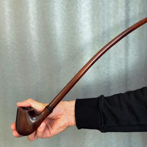 Könnte beinhalten: Eine dunkelbraune Pfeife mit einem langen, gebogenen Stiel. Die Pfeife hat einen abgerundeten Kopf und einen schlanken Stiel. Der Stiel wird in einer Hand gehalten, mit einem schwarzen Ärmel im Hintergrund. Die Pfeife scheint aus Holz zu sein.