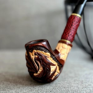 May include: A tobacco pipe featuring a dark brown wooden bowl intricately carved with a dragon motif. The shank displays a lighter wood section with a textured pattern, followed by a red and black braided cord wrap and a black stem with gold trim.