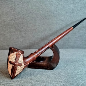 Puede incluir: Una pipa de tabaco de madera con un tallo largo y una boquilla curva. El cuenco presenta un diseño de escudo tallado con una espada y una cruz. La pipa descansa sobre un soporte de madera, mostrando su intrincado diseño.