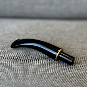 以下が含まれることがあります： 黒いパイプの吸い口で、曲線的なデザインと金色の金属リングが付いています。吸い口は光沢があり滑らかで、ステムは灰色のテクスチャのある表面に置かれています。これは喫煙パイプの交換部品です。