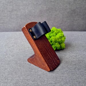 May include: A dark brown wooden stand with a slanted top surface featuring a black leather strap secured by two small brass-coloured metal rivets. The stand has a solid, angled base and a smooth, polished wood grain finish throughout.