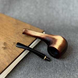 May include: A tobacco pipe with a polished dark brown wooden bowl featuring lighter wood grain highlights and a detachable black stem with a thin gold-coloured metal ring at the connection point.