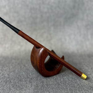 Puede incluir: Una pipa de madera con boquilla negra y un acento dorado en el extremo. La pipa descansa en un soporte de madera a juego. La pipa y el soporte son de madera marrón oscuro.
