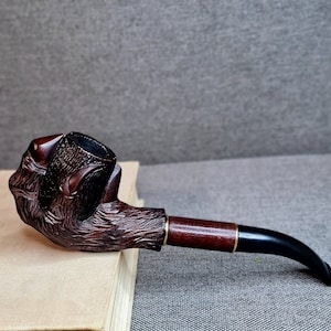 May include: A wooden smoking pipe carved in the shape of a hand holding a bowl. The pipe is brown and has a black stem with a gold band.