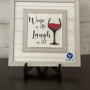 以下が含まれることがあります： 白い木製の額縁に入ったサインで、赤いワイングラスと「Wine a little Laugh a lot」というテキストが白い背景に書かれています。
