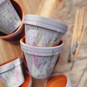 May include: Several lavender-painted flower pots with orange interiors are arranged on a wooden surface. The pots are decorated with lavender and green floral designs. Paintbrushes are visible in the background, suggesting a craft or DIY project.