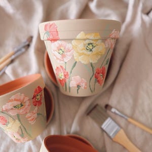 May include: Tan-colored ceramic flower pots with hand-painted poppy designs in shades of pink, yellow, and red. The pots have matching saucers and are surrounded by paintbrushes, suggesting a craft or gardening theme.
