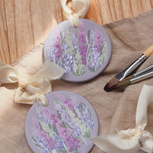 Pode incluir: Enfeites redondos de cerâmica cor lavanda com desenhos florais pintados à mão em tons de roxo, rosa e verde. Cada enfeite é amarrado com uma fita creme. Pincéis são visíveis, sugerindo um tema artístico.