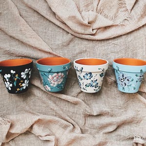 May include: Four terracotta flower pots with hand-painted floral designs. The pots are arranged in a row on a light brown fabric background. The designs include white flowers on a black pot, pink flowers on a blue pot, blue flowers on a white pot, and purple flowers on a blue pot.