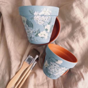 May include: Two pale blue flower pots with white floral patterns and matching saucers. The pots have an orange interior. Paintbrushes are placed nearby, suggesting a handmade or DIY theme. The background is a neutral fabric.