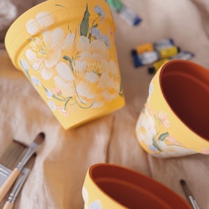 May include: Three yellow flower pots with hand-painted floral designs. The pots have a terracotta interior and are decorated with white and blue flowers. Paint tubes and brushes are nearby.